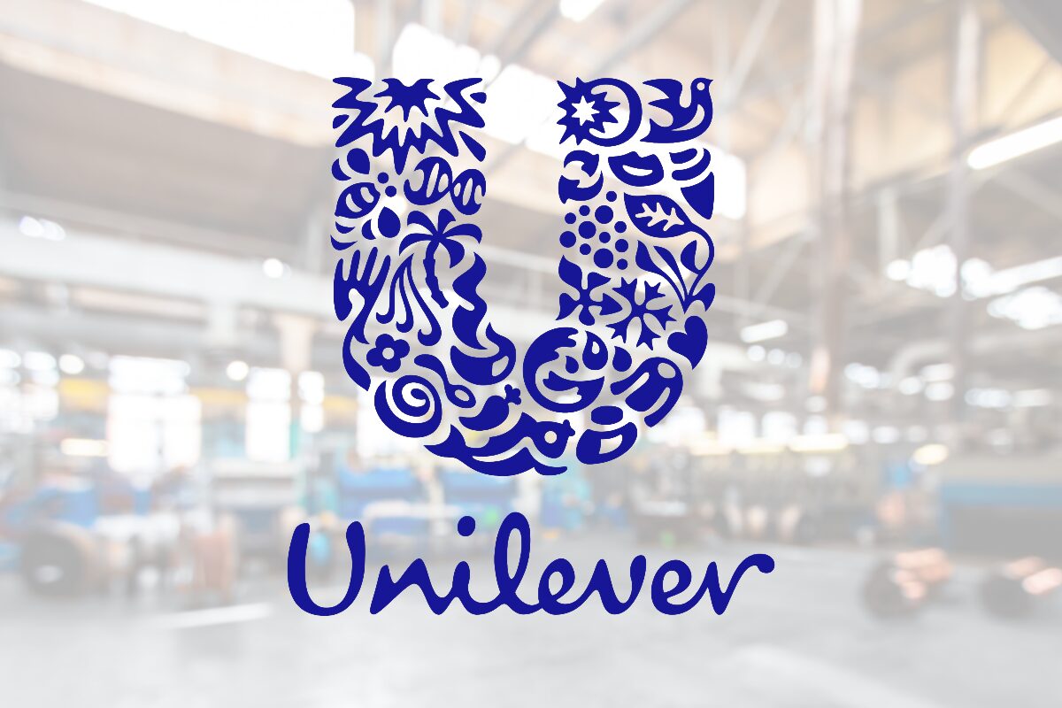 Blue Unilever logo: a decorative U made of floral shapes with the word Unilever beneath, shown on a glass surface in a blurred industrial background.