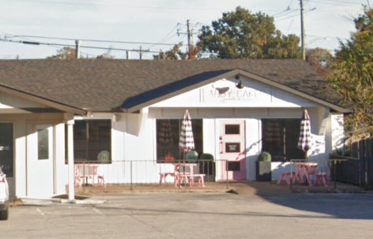 Small white cafe with pink door and outdoor seating, pink tables, and striped umbrellas outside the entrance.