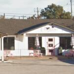 Small white cafe with pink door and outdoor seating, pink tables, and striped umbrellas outside the entrance.