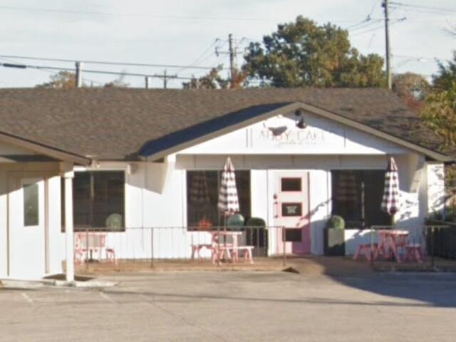 Small white cafe with pink door and outdoor seating, pink tables, and striped umbrellas outside the entrance.