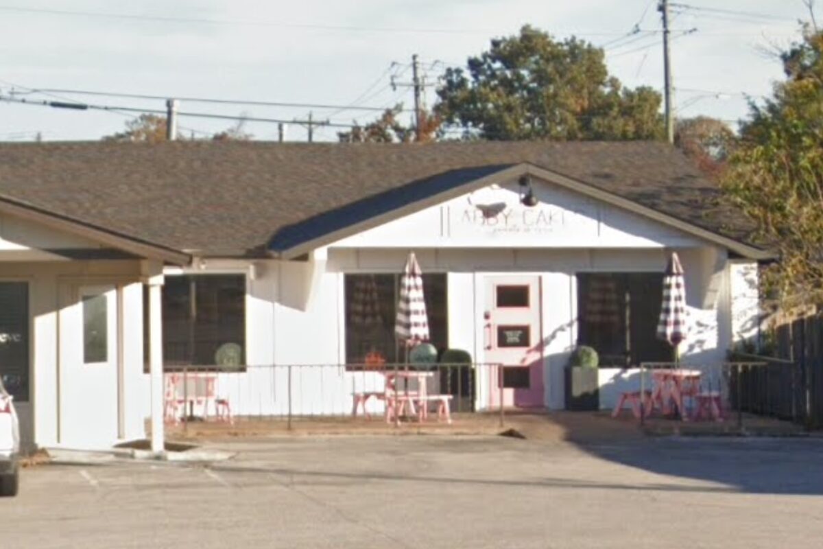 Small white cafe with pink door and outdoor seating, pink tables, and striped umbrellas outside the entrance.
