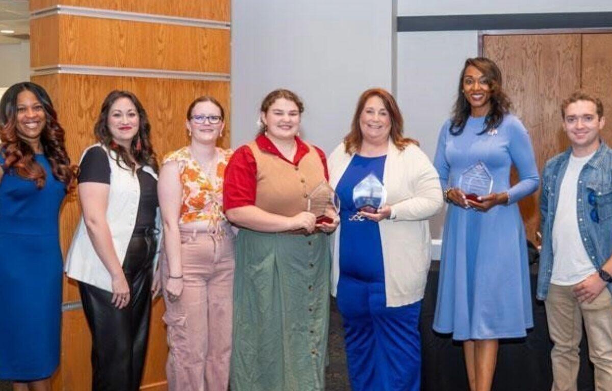 Group of eight people standing in a row, smiling, each holding awards after a ceremony in a modern lobby.