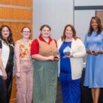 Group of eight people standing in a row, smiling, each holding awards after a ceremony in a modern lobby.