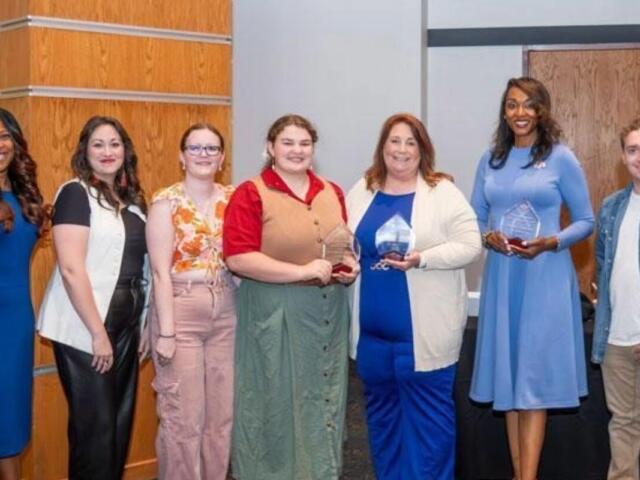 Group of eight people standing in a row, smiling, each holding awards after a ceremony in a modern lobby.