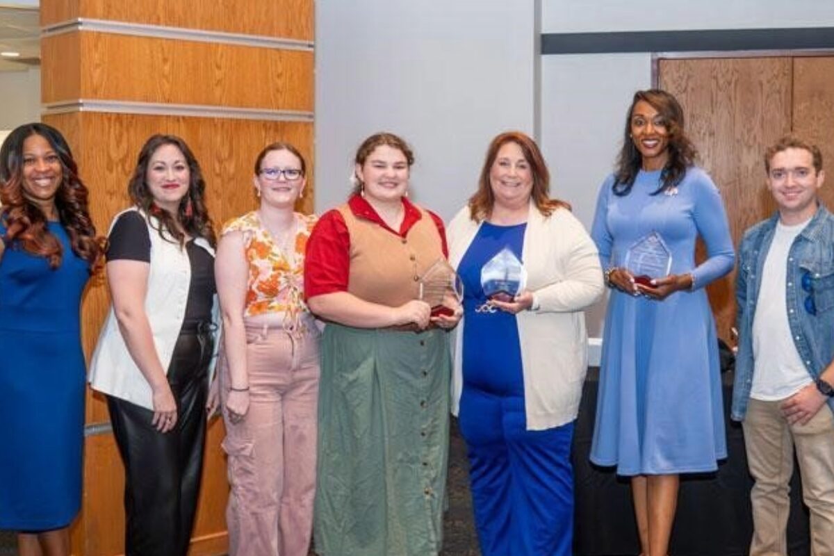 Group of eight people standing in a row, smiling, each holding awards after a ceremony in a modern lobby.