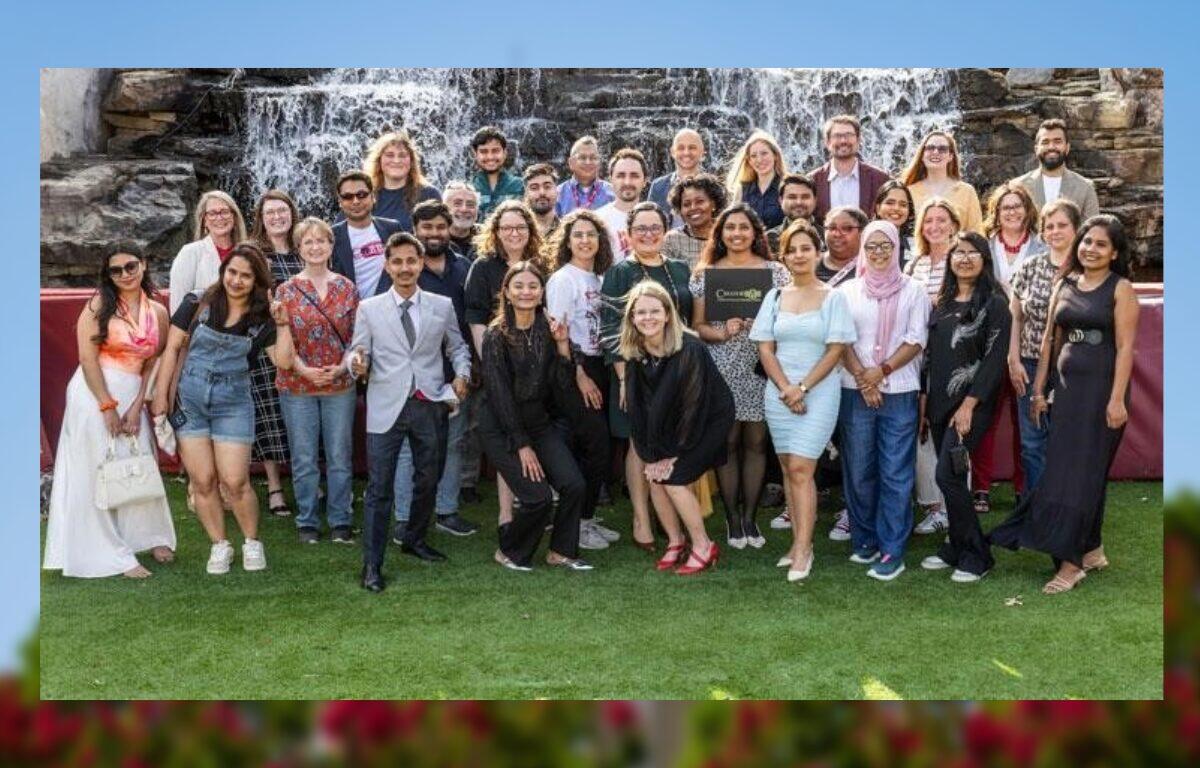 Diverse group of people posing for a group photo on a grassy outdoor area with a waterfall backdrop behind them.