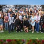 Diverse group of people posing for a group photo on a grassy outdoor area with a waterfall backdrop behind them.