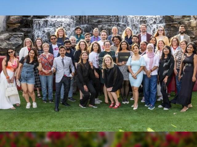 Diverse group of people posing for a group photo on a grassy outdoor area with a waterfall backdrop behind them.