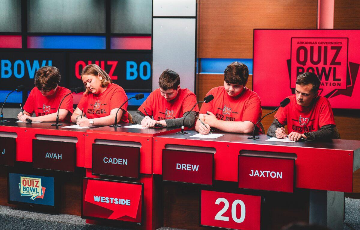 Five students in red shirts sit at a quiz bowl panel with microphones; nameplates read AVAH, CADEN, DREW, JAXTON; score 20 on a red panel.