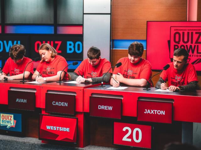 Five students in red shirts sit at a quiz bowl panel with microphones; nameplates read AVAH, CADEN, DREW, JAXTON; score 20 on a red panel.