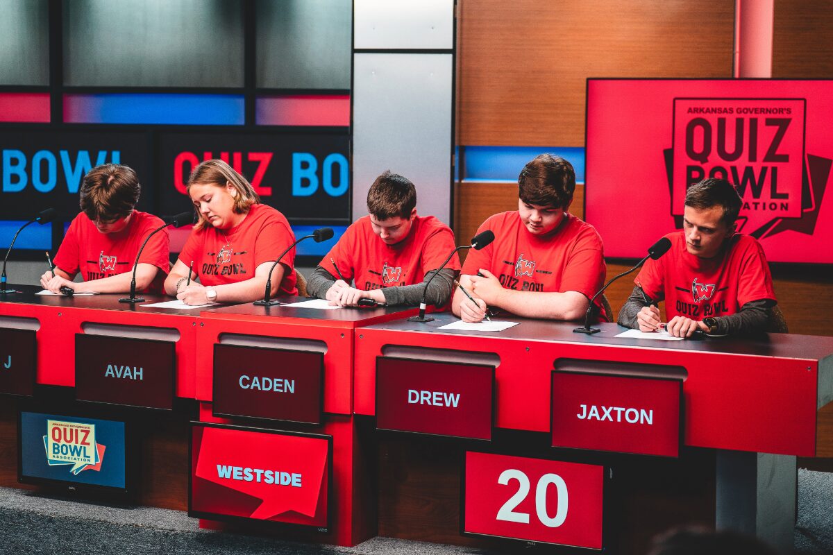 Five students in red shirts sit at a quiz bowl panel with microphones; nameplates read AVAH, CADEN, DREW, JAXTON; score 20 on a red panel.