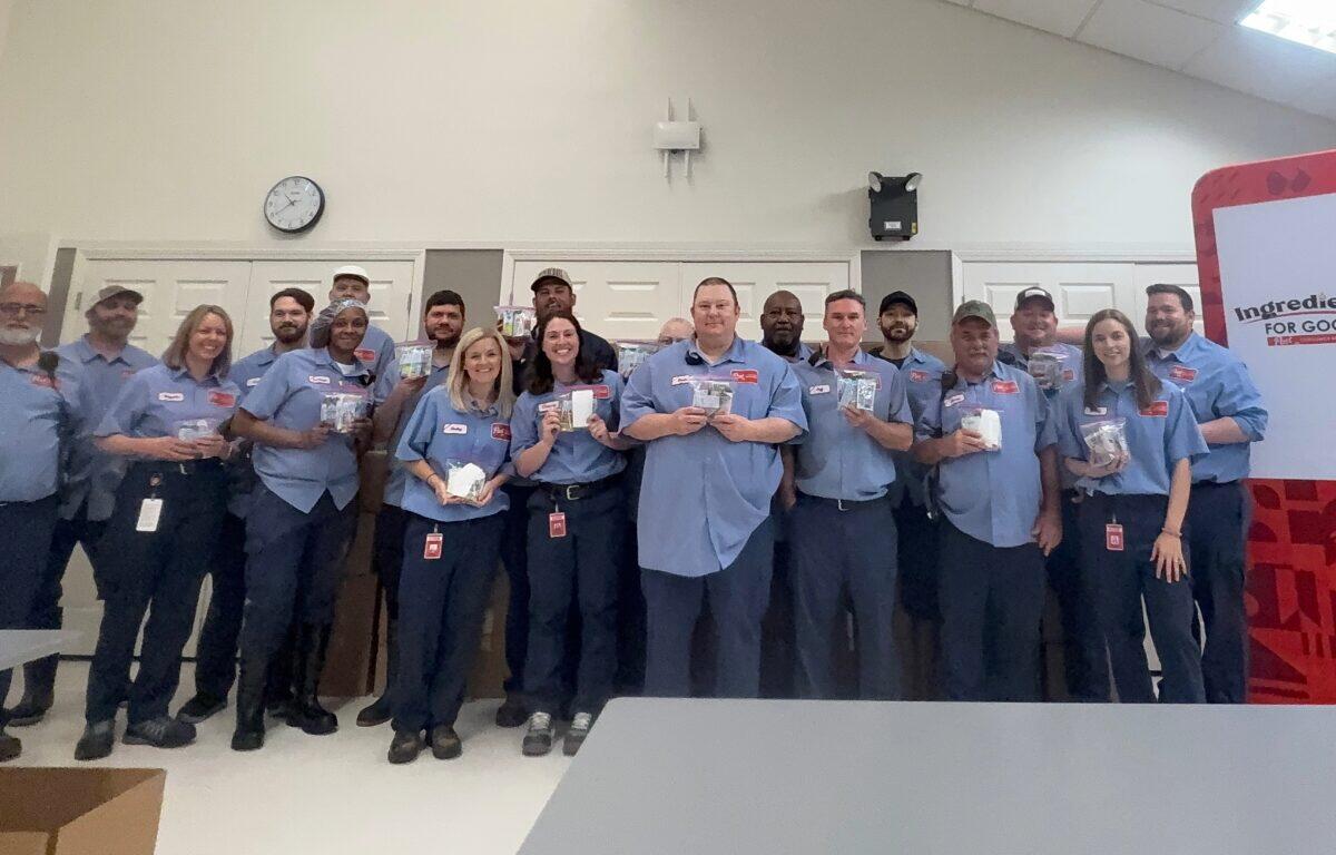 Group of coworkers in blue shirts posing for a photo in a break room, each holding small items or samples, smiling at the camera.