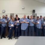 Group of coworkers in blue shirts posing for a photo in a break room, each holding small items or samples, smiling at the camera.