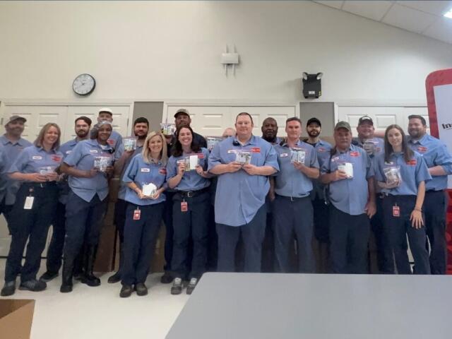 Group of coworkers in blue shirts posing for a photo in a break room, each holding small items or samples, smiling at the camera.