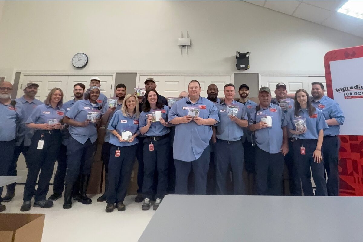 Group of coworkers in blue shirts posing for a photo in a break room, each holding small items or samples, smiling at the camera.