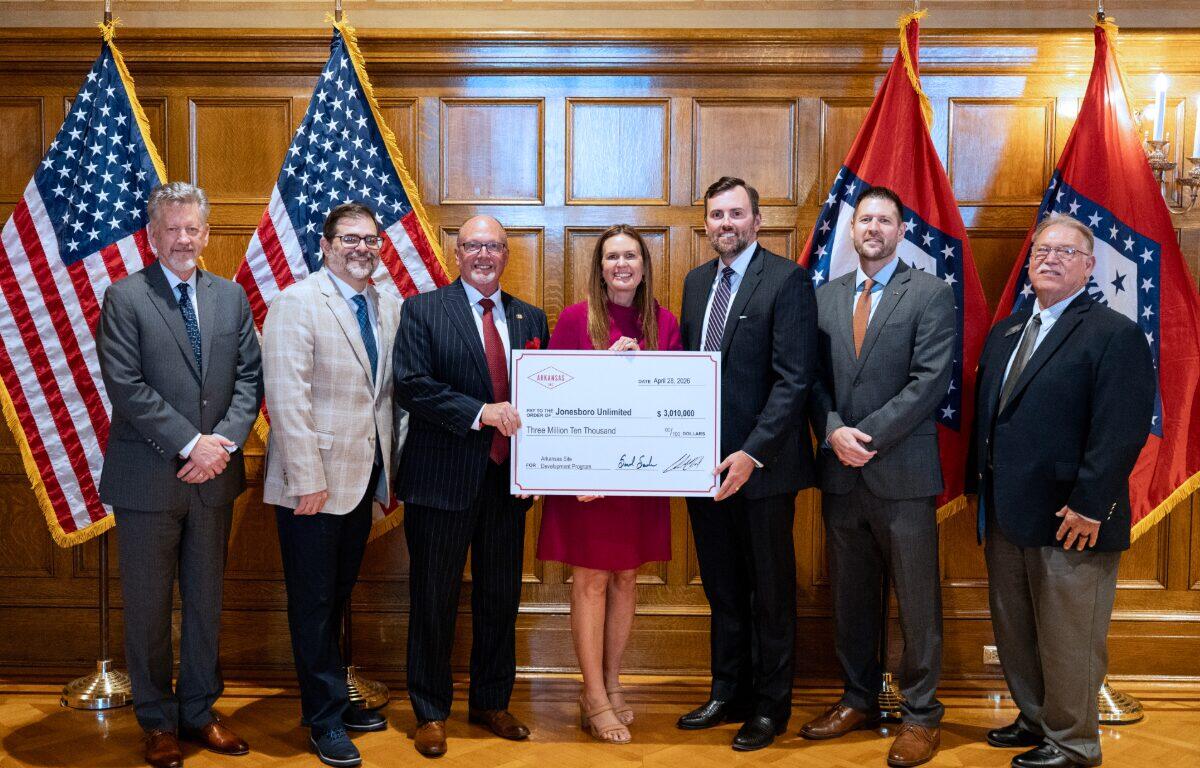 Seven professionals in business attire pose with a large ceremonial check in a wood-paneled room, American flags on either side of the group.