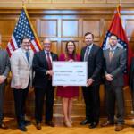 Seven professionals in business attire pose with a large ceremonial check in a wood-paneled room, American flags on either side of the group.