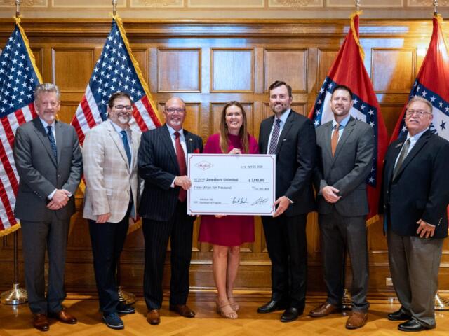 Seven professionals in business attire pose with a large ceremonial check in a wood-paneled room, American flags on either side of the group.