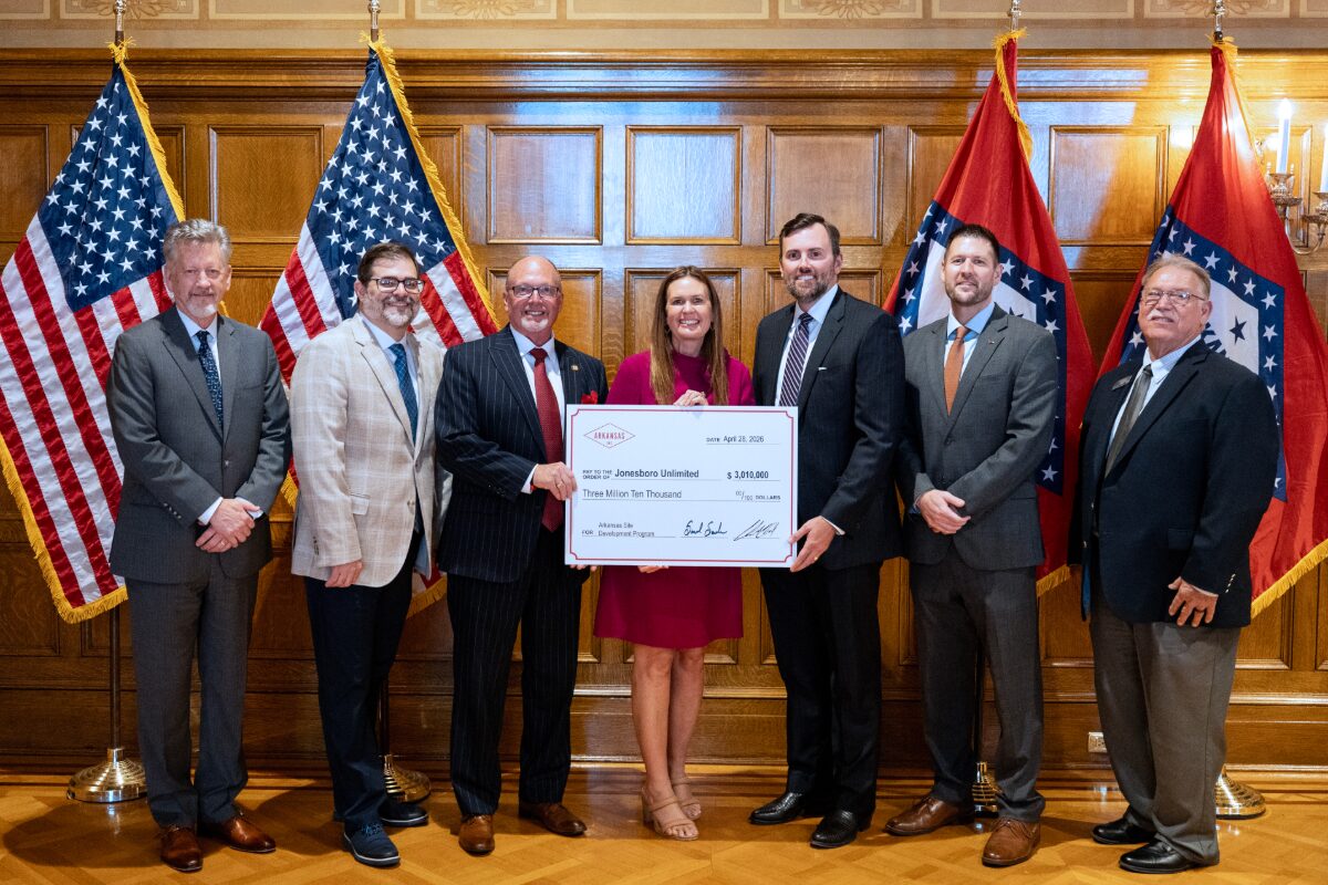 Seven professionals in business attire pose with a large ceremonial check in a wood-paneled room, American flags on either side of the group.