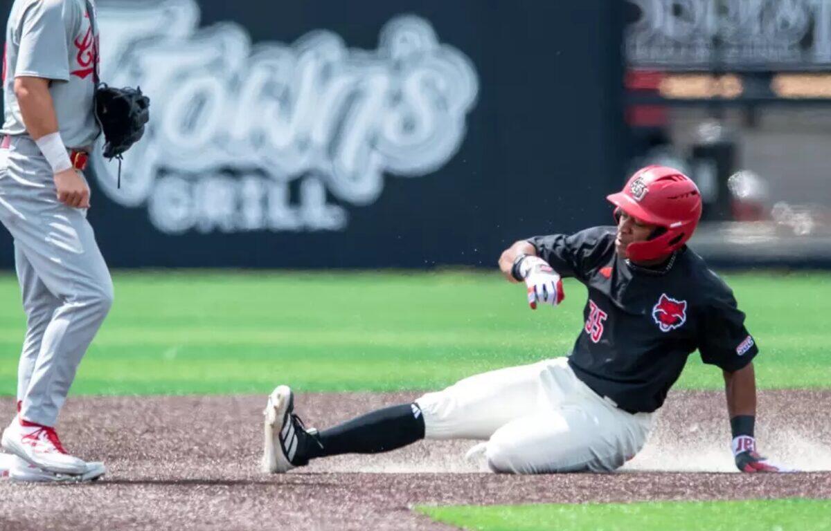 Baseball player in black jersey slides into a base, kicking up dust as a fielder stands nearby in gray uniform.