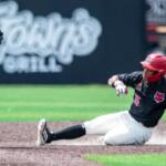 Baseball player in black jersey slides into a base, kicking up dust as a fielder stands nearby in gray uniform.