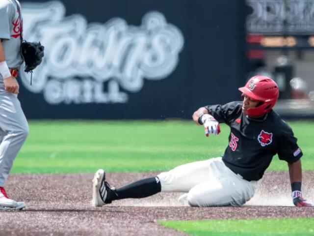 Baseball player in black jersey slides into a base, kicking up dust as a fielder stands nearby in gray uniform.