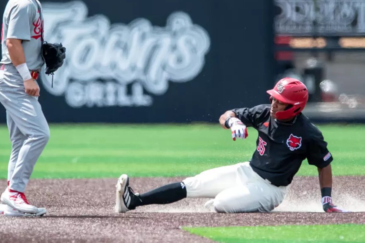 Baseball player in black jersey slides into a base, kicking up dust as a fielder stands nearby in gray uniform.
