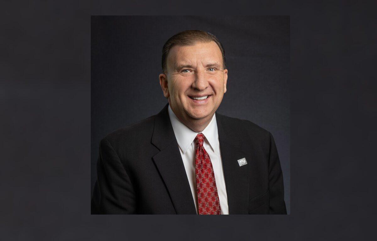 Smiling man in a dark suit, white shirt, and red patterned tie, wearing a lapel pin, posed against a dark backdrop for a professional portrait