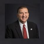 Smiling man in a dark suit, white shirt, and red patterned tie, wearing a lapel pin, posed against a dark backdrop for a professional portrait