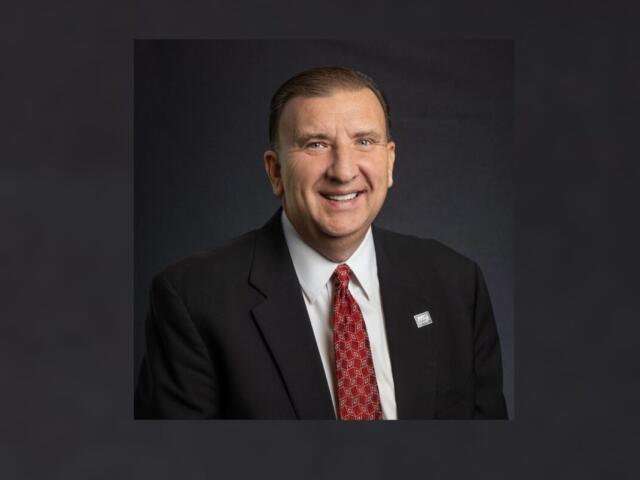 Smiling man in a dark suit, white shirt, and red patterned tie, wearing a lapel pin, posed against a dark backdrop for a professional portrait