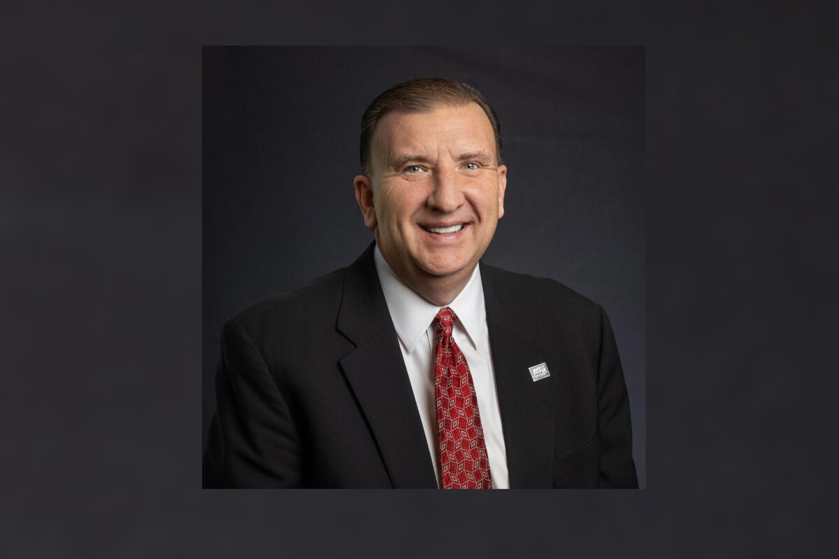 Smiling man in a dark suit, white shirt, and red patterned tie, wearing a lapel pin, posed against a dark backdrop for a professional portrait