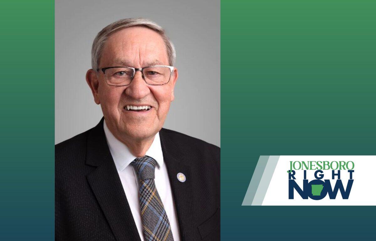 Smiling older man in a dark suit, white shirt, and patterned tie, wearing glasses, against a gray backdrop with a 'Jonesboro Right Now' logo on the right.