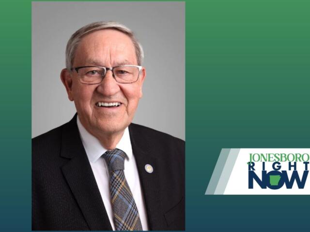 Smiling older man in a dark suit, white shirt, and patterned tie, wearing glasses, against a gray backdrop with a 'Jonesboro Right Now' logo on the right.