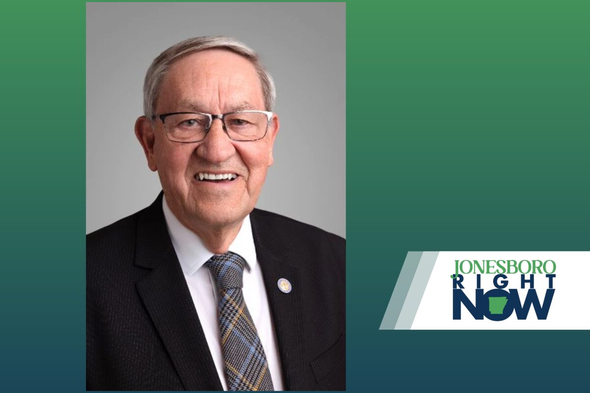 Smiling older man in a dark suit, white shirt, and patterned tie, wearing glasses, against a gray backdrop with a 'Jonesboro Right Now' logo on the right.