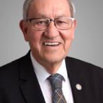 Portrait of an elderly man in a dark suit and patterned tie, wearing glasses, smiling at the camera.
