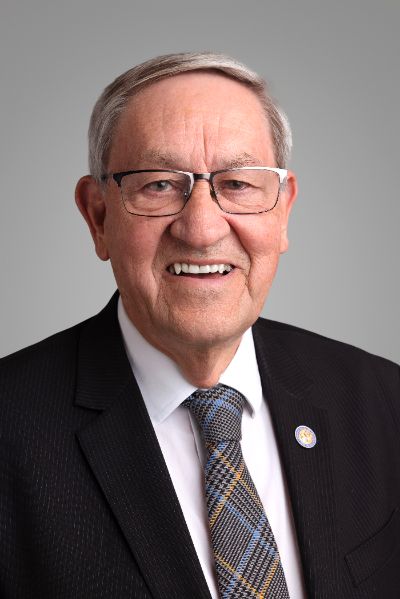 Portrait of an elderly man in a dark suit and patterned tie, wearing glasses, smiling at the camera.