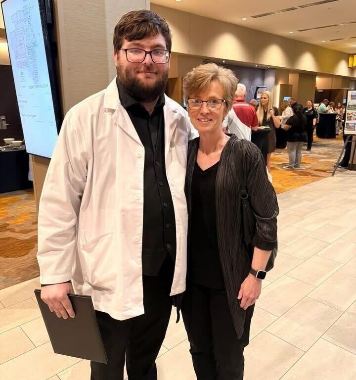 A man in a white lab coat and black outfit stands beside a woman in a black cardigan at a conference, both smiling for the camera with a clipboard in his hand and other attendees in the background.