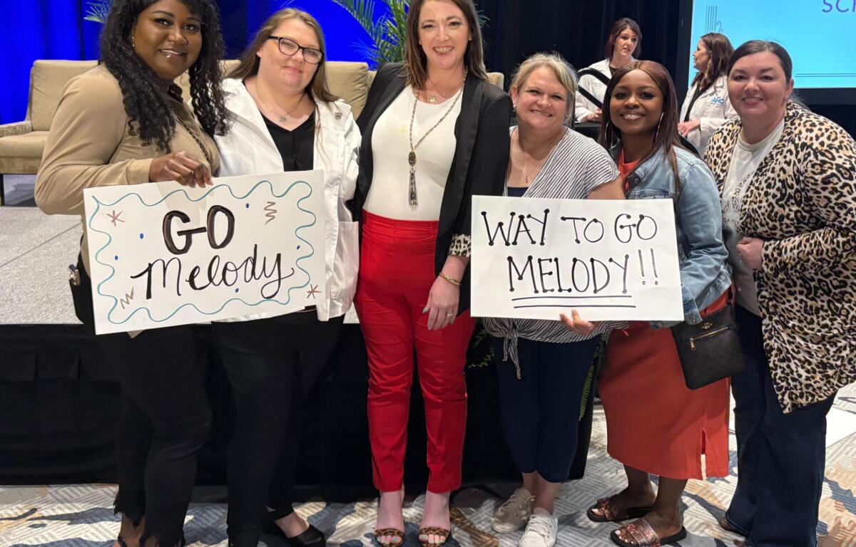 Group of seven women on a stage, smiling; two hold signs reading 'GO Melody' and 'WAY TO GO MELODY!!'