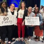 Group of seven women on a stage, smiling; two hold signs reading 'GO Melody' and 'WAY TO GO MELODY!!'