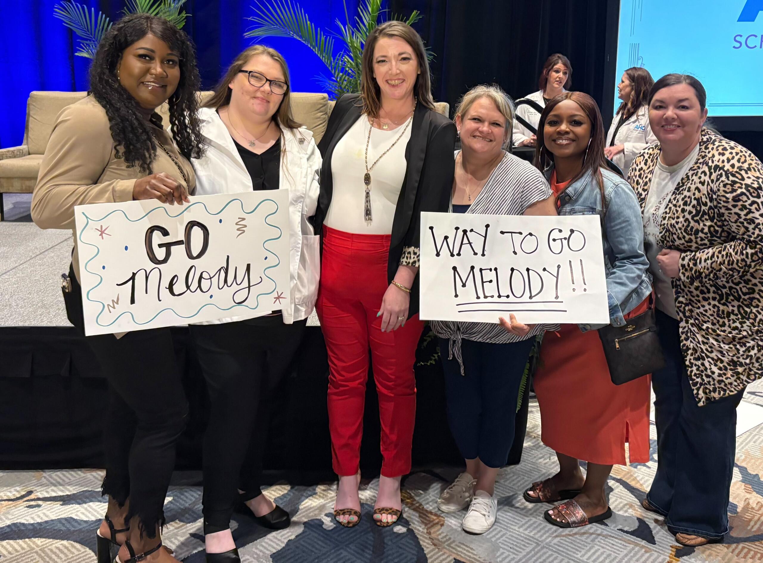 Group of seven women on a stage, smiling; two hold signs reading 'GO Melody' and 'WAY TO GO MELODY!!'