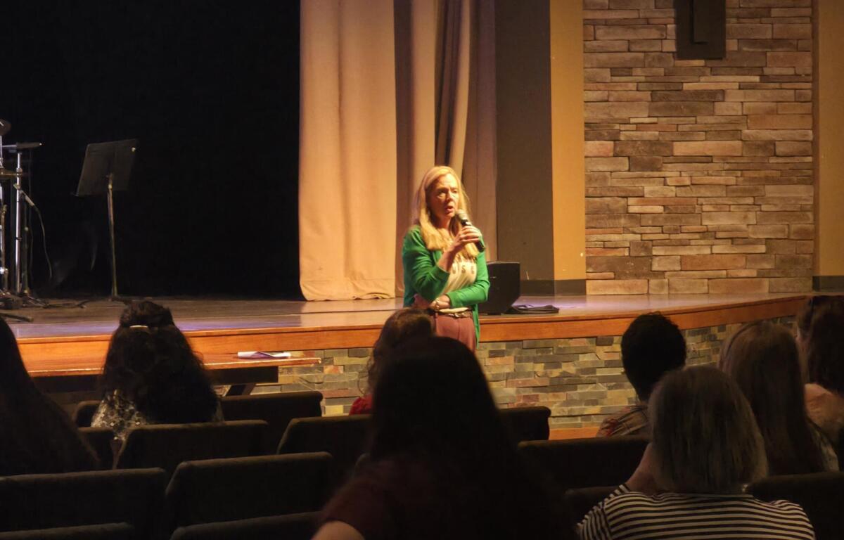 Woman in a green cardigan on stage holding a microphone, speaking to an audience in a theater with brick wall backdrop.