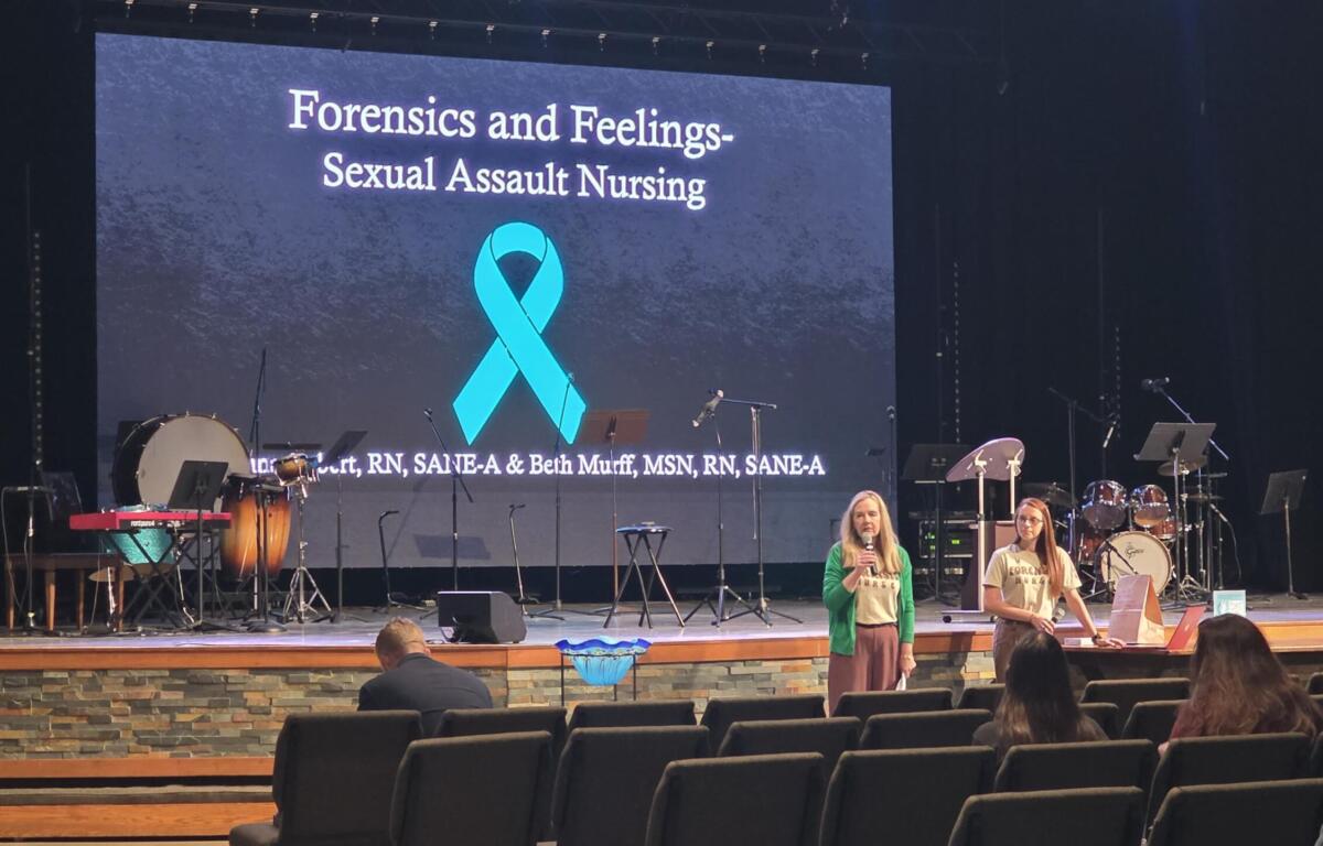 Stage with a large screen showing 'Forensics and Feelings - Sexual Assault Nursing' and a teal ribbon, two women speaking at microphones.