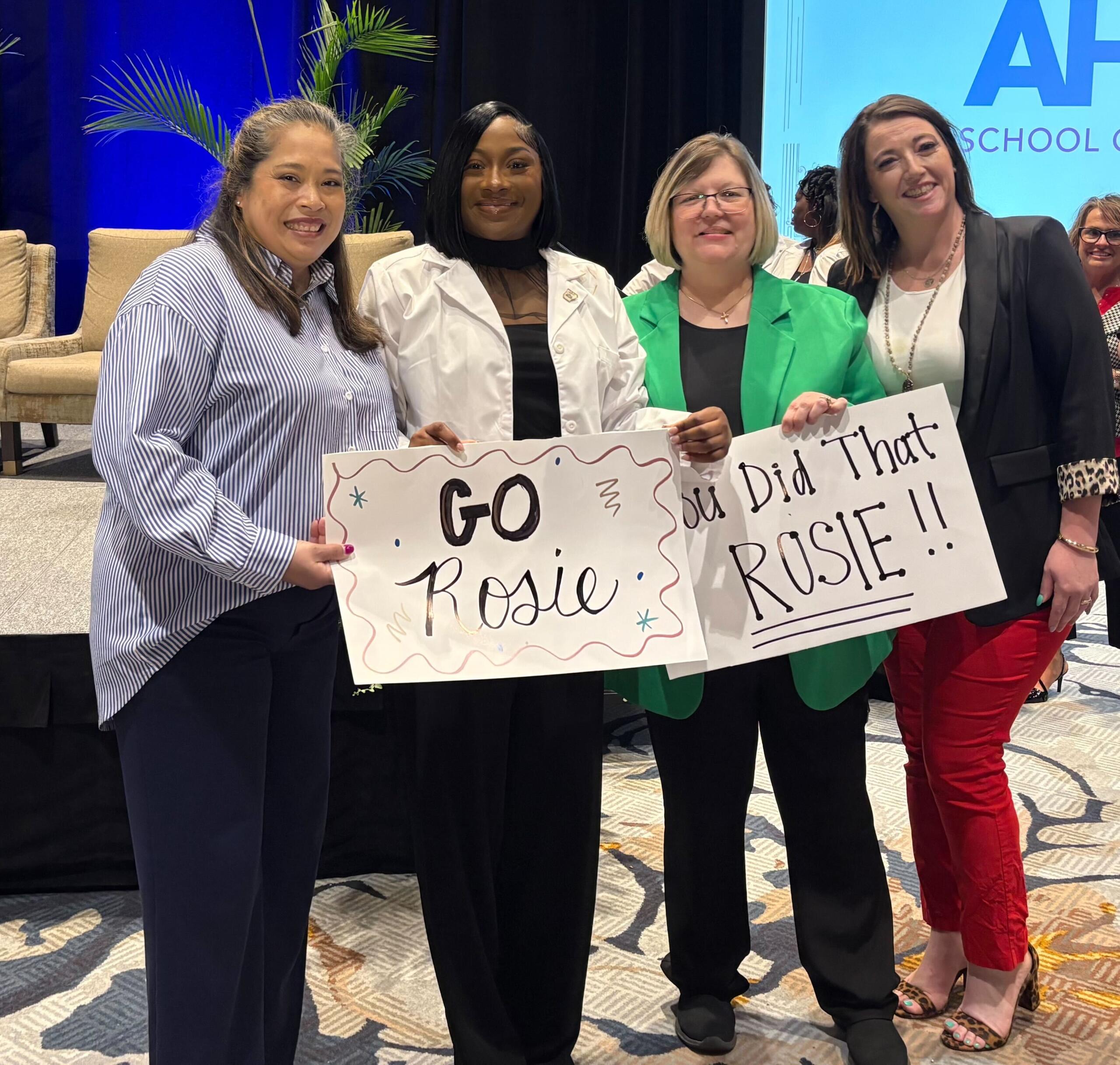 Four women smiling on a stage, holding signs that read 'GO Rosie' and 'Did That Rosie!!' in support.
