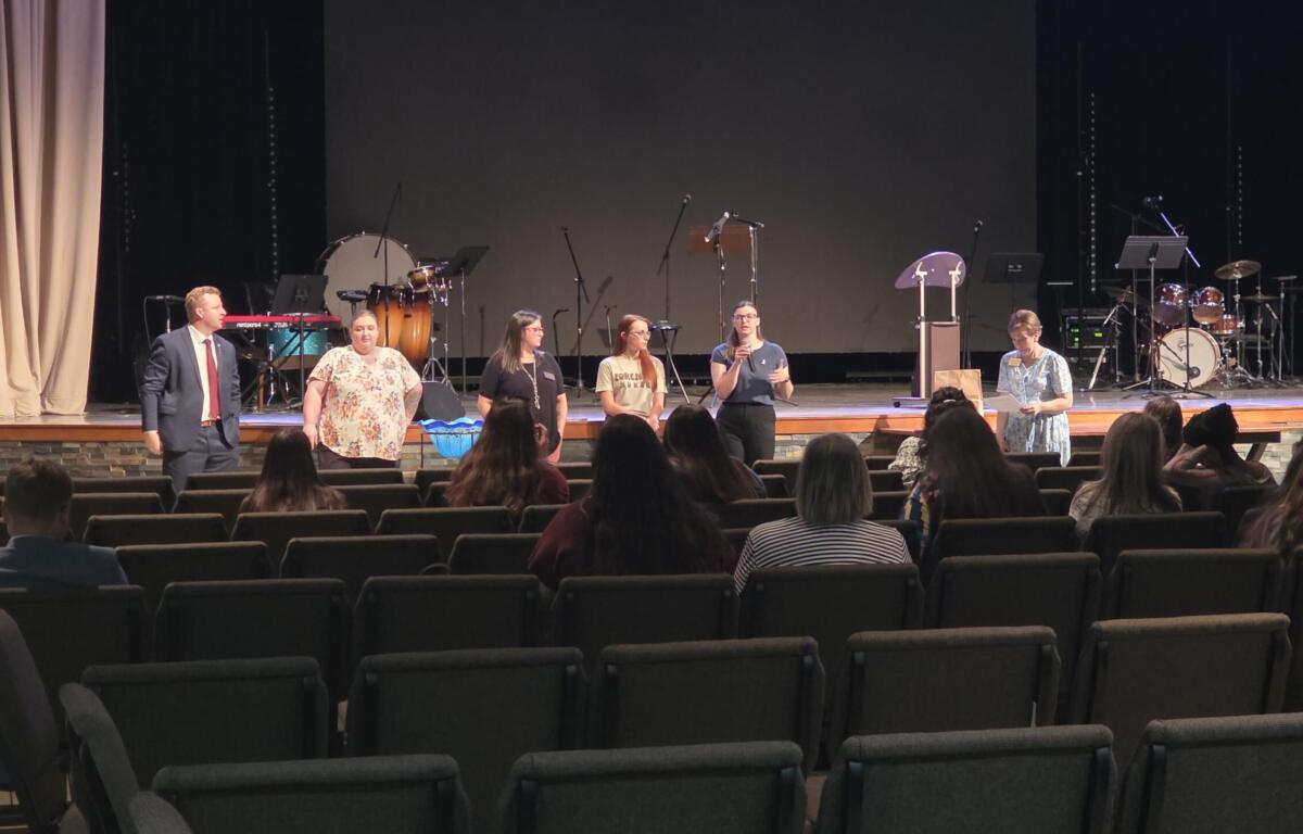 Man in a suit and five women stand on a stage addressing an audience; musical instruments are visible in the background.