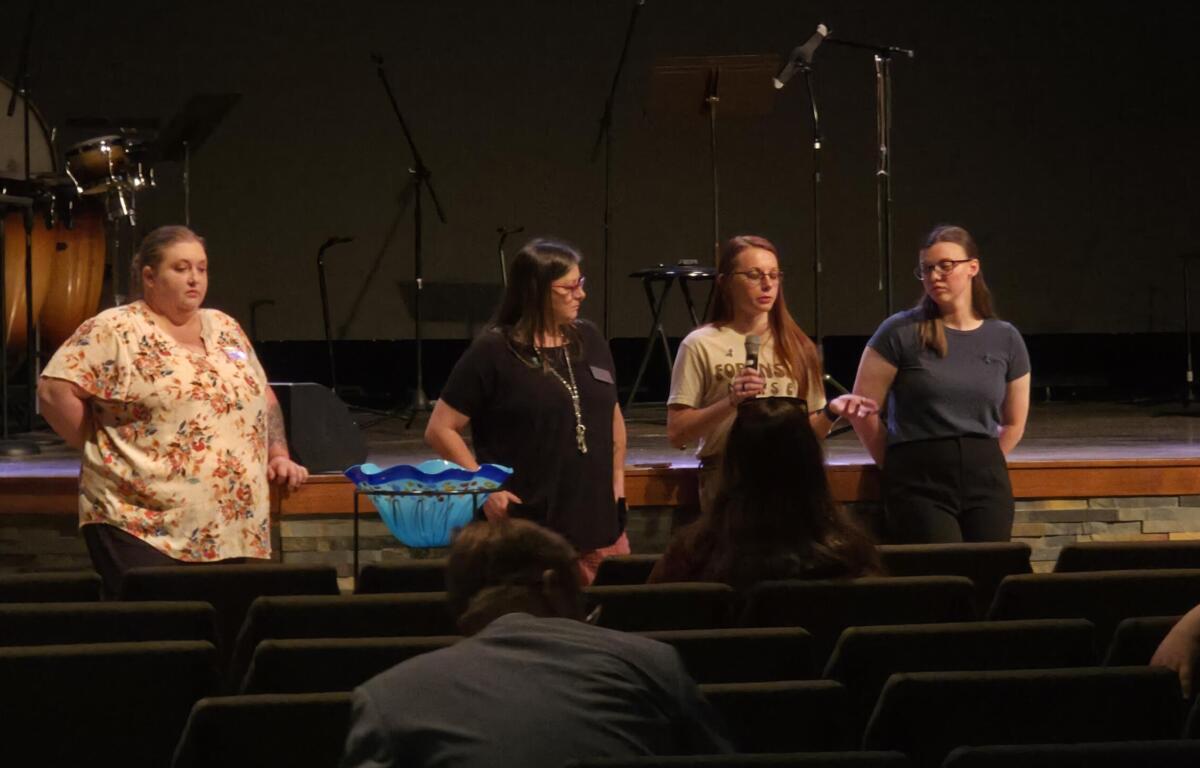 Four women stand on a dimly lit stage; one holds a microphone while three others listen, with musical equipment in the background and audience seating in front.