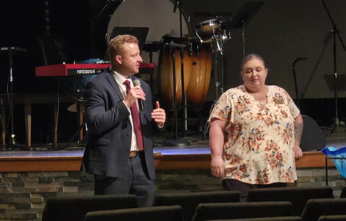 Man in a dark suit and red tie speaks into a handheld microphone on a stage beside a woman in a floral blouse, with musical instruments in the background.