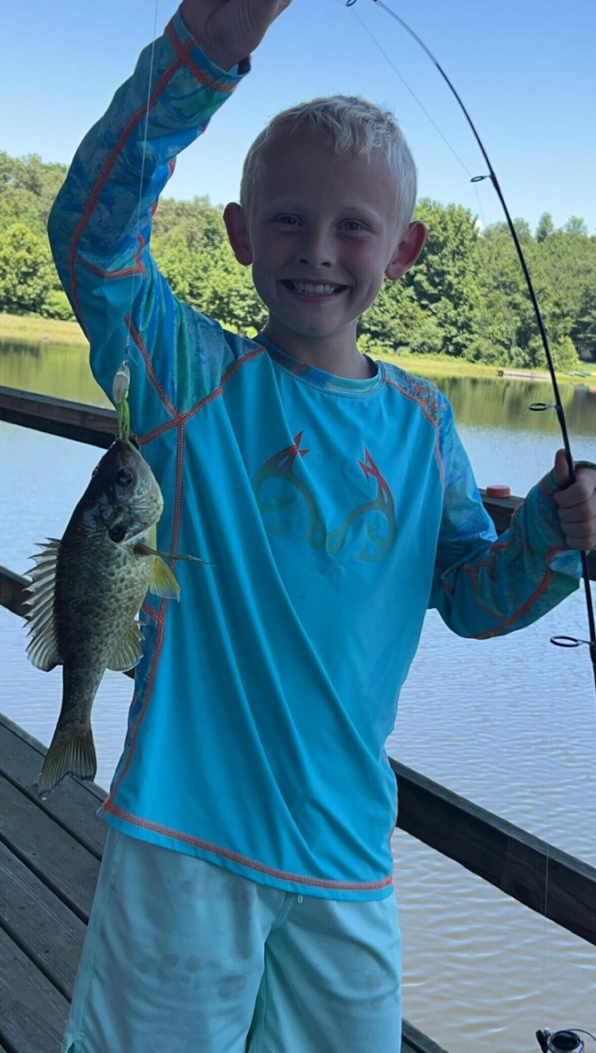 Smiling boy on a wooden dock holding up a small fish he caught, with a fishing rod in the other hand and a lake and trees in the background.
