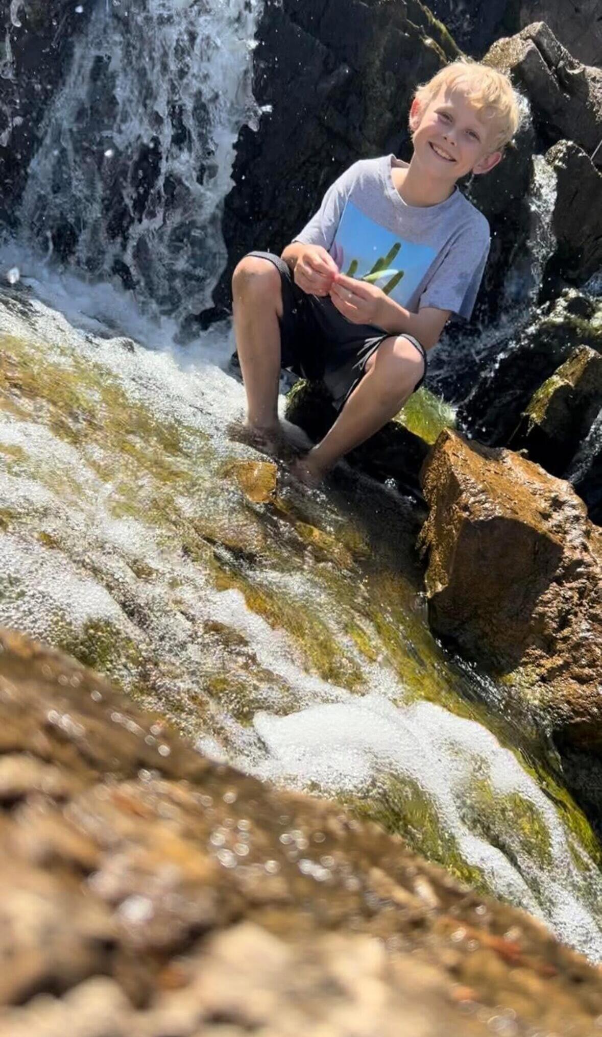 Smiling boy sits on wet rocks beside a small waterfall, feet in the rushing water.