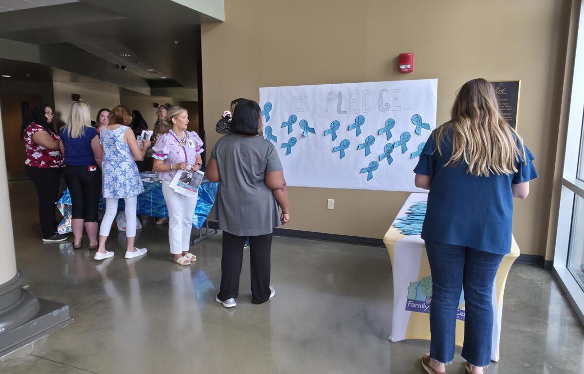 Group of people gathered in a hallway near a large poster that reads 'I PLEDGE...' with blue ribbons arranged on it, likely a pledge event area.