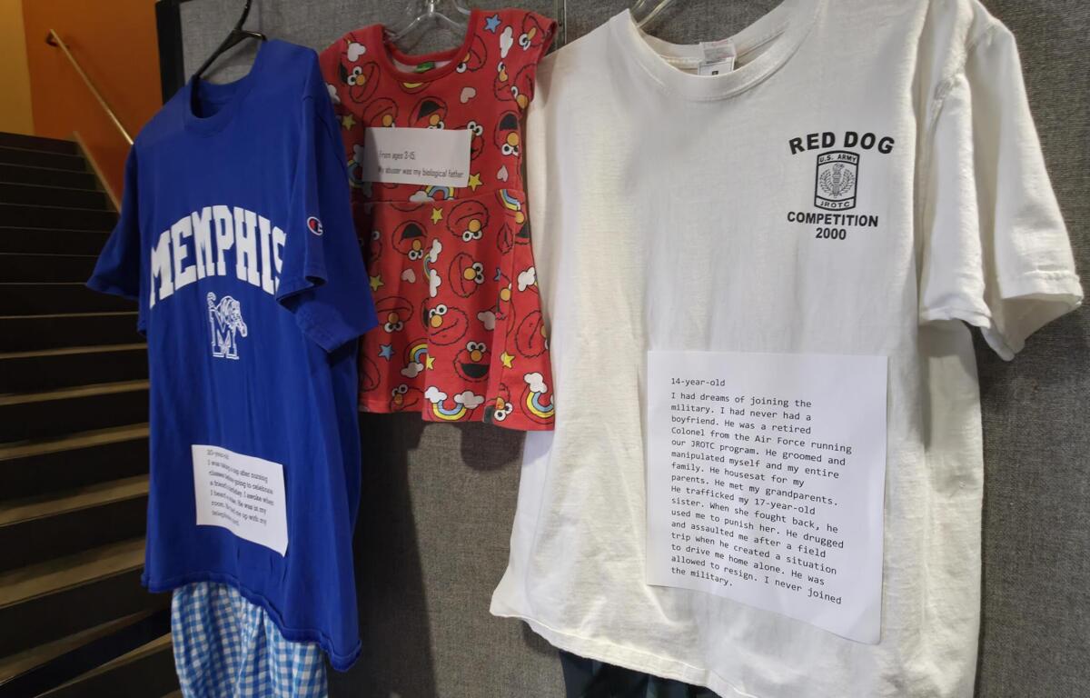 Blue Memphis T-shirt on a hanger next to a red Elmo-patterned shirt and a white "Red Dog" t-shirt with a paper label in front of a gray wall.